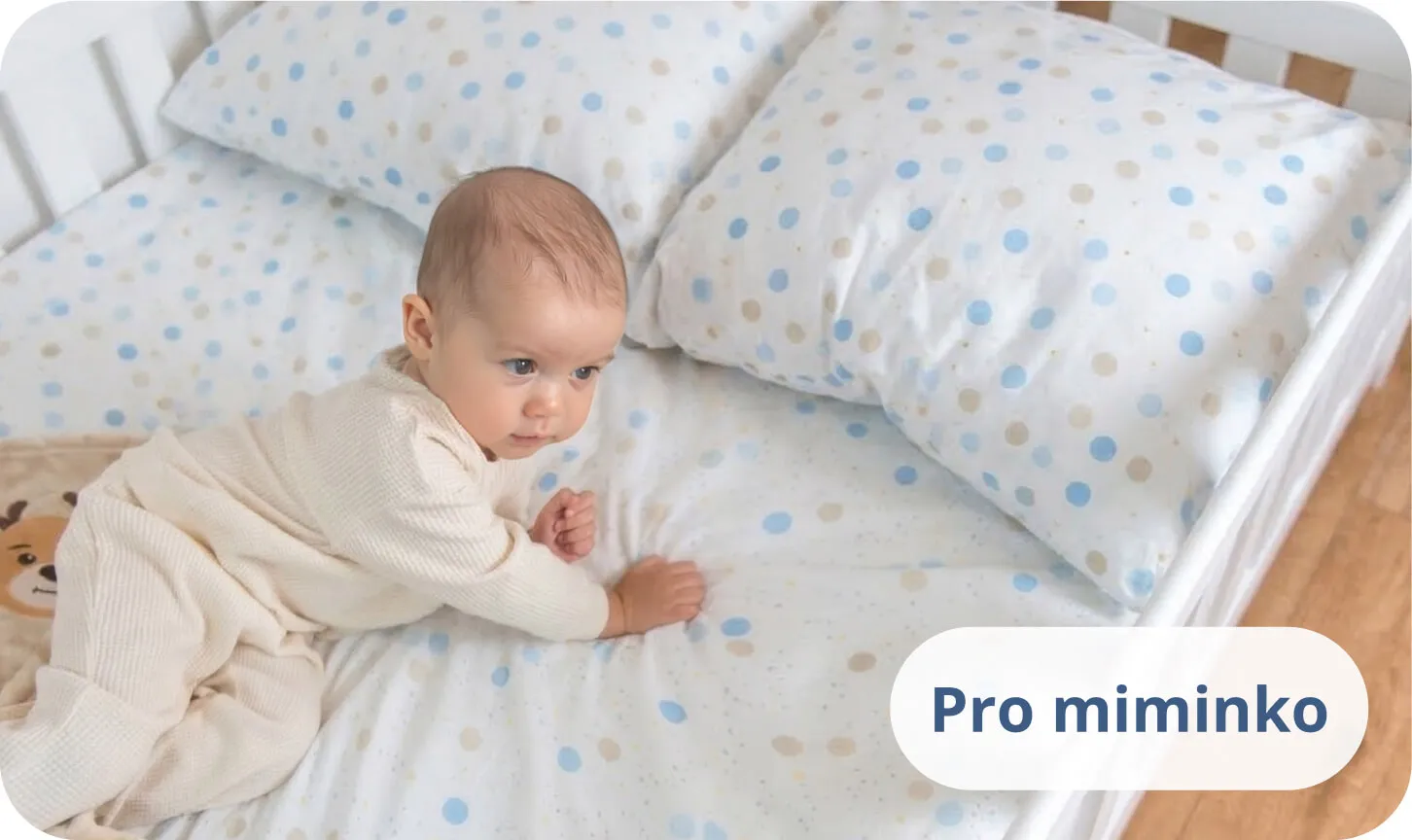 Miminko ležící na bříšku na bílé posteli v povlečení s modrými puntíky.